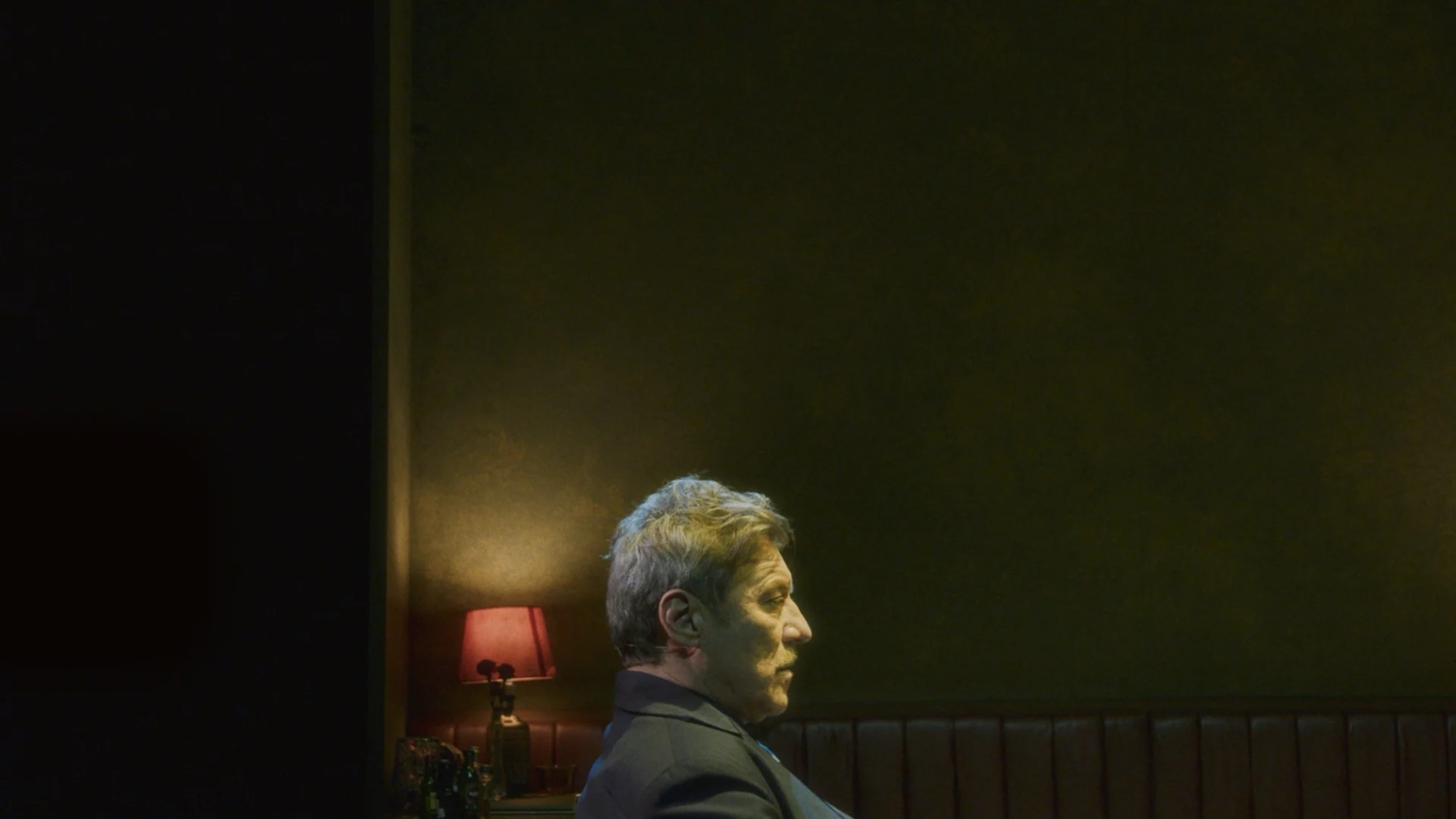 Grey-haired man in profile in a dimly lit olive-green room, a red table lamp glowing behind him