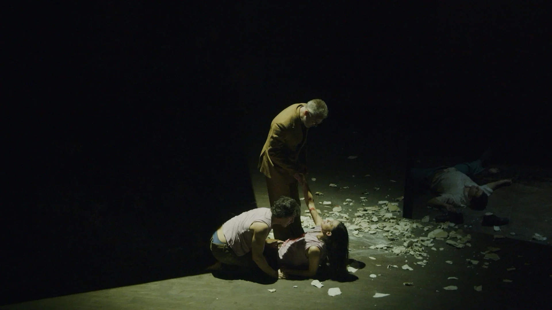 Stage scene in a narrow pool of light — a standing figure reaches down to two people on a floor strewn with torn paper